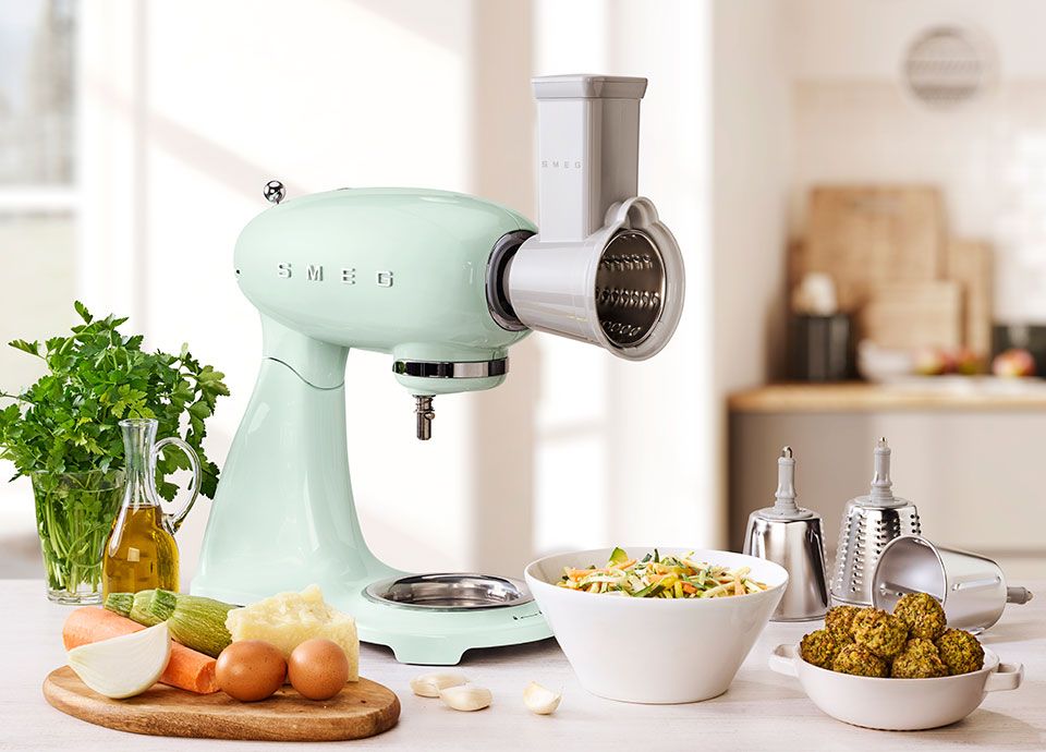 A green smeg food mixer with a vegetable cutter accessory attached to the front, and beneath it, is a bowl of freshly cut vegetables such as carrots, peppers and cucumbers