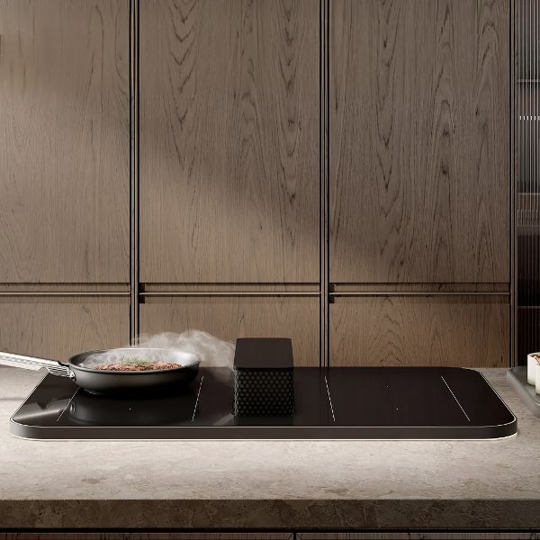 Black glass induction hob with integrated downdraft extractor, cooking pan in use, installed in a kitchen island with wood panel background.