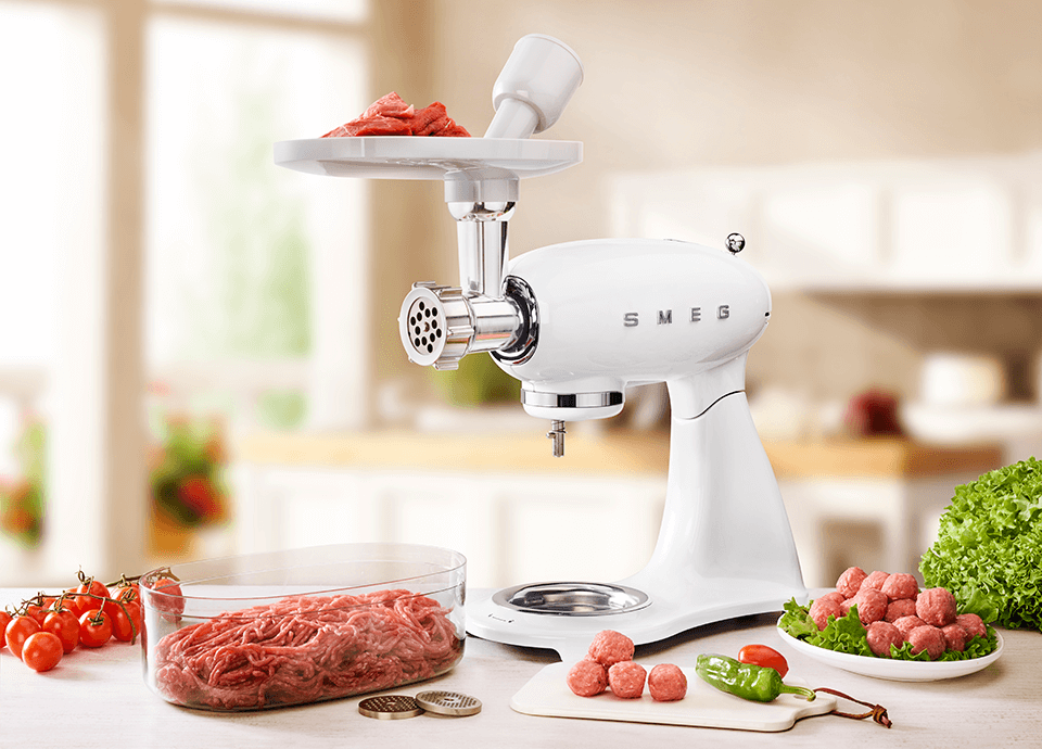 A cream retro Smeg food mixer on a kitchen worktop surrounded various meats being prepared for a BBQ. Attached to the front is a food grinder ready to prepare meat into a fine mince